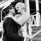 Ein Paar in eleganter Kleidung, sie trägt ein glitzerndes Kleid, er eine Matrosenmütze, steht vor einer Veranda mit Geländer.