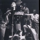 Eine Frau hält einen Mann am Boden, umgeben von einem Kamerateam mit großen Scheinwerfern. Retro Stil.