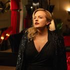 Eine Frau in schwarzem, glitzerndem Kleid steht in einem stilvollen Raum mit rotem Samt und gedämpfter Beleuchtung.