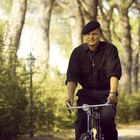 Eine ältere Person trägt eine schwarze Kleidung mit einem Kreuz und fährt auf einem Fahrrad durch einen sonnigen Waldweg.