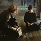 Es gibt keine sichtbaren Texte. Eine Frau und ein Junge sitzen nah beieinander auf einem Holzboden in einem dunklen Raum.