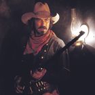 Ein Cowboy mit Hut und rotem Halstuch hält ein Gewehr vor einer leuchtenden Öllampe in einem dunklen Raum.