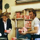 Ein Mann in einem Anzug und ein anderer in einem T-Shirt packen rote Pakete aus einer beschädigten Wand.