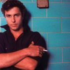 Eine Person lehnt an einer türkisfarbenen Kachelwand und hält eine Zigarette.