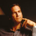 In sanftem Licht sitzt ein nachdenklicher Mann, die Hand am Kinn, mit einem grauen Hemd und einer Armbanduhr.