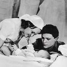 Eine Frau und ein Mann betrachten liebevoll ein Baby, das auf einem Bett liegt.