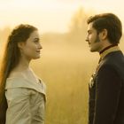 Eine Frau und ein Mann stehen sich in einem Feld gegenüber, bei warmem, goldenem Licht im Hintergrund.