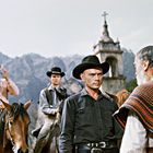 Männer in Cowboy-Kleidung, zwei auf Pferden, blicken ernst drein; Kirchturm im Hintergrund.