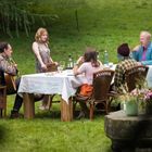 Sechs Personen sitzen im Freien an einem Tisch. Eine Person spielt Gitarre, während die anderen essen und trinken. Grünen Rasen umgibt sie.