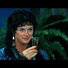 Eine Frau mit dunklen Haaren hält ein Glas in der Hand. Sie trägt Ohrringe. Im Hintergrund sind Pflanzen sichtbar.