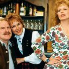 Keine Texte sichtbar. Drei lächelnde Personen in traditioneller Kleidung vor einem Schrank mit Gläsern und Flaschen.