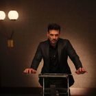 Ein Mann in einem dunklen Anzug steht ernst an einem durchsichtigen Pult in einem schummrig beleuchteten Raum. Links eine Wandleuchte.