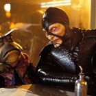 Ein Alien und ein Mensch in schwarzem Kostüm sitzen nebeneinander an einem Tisch in einer futuristischen Umgebung.