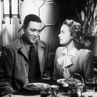 Zwei Personen in eleganter Kleidung sitzen an einem Tisch in einem stilvollen Raum und schauen sich an.