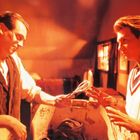 Es gibt keinen sichtbaren Text. Zwei Männer in einem orange beleuchteten Raum; der eine reicht dem anderen eine Schere.