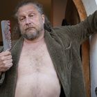 „Kayser“ auf einem Türschild. Ein Mann, der müde aussieht, hält ein blutiges Messer. Er trägt einen offenen Bademantel.