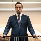 Ein Mann im Anzug steht hinter einem Geländer, in einem modernen und hellen Innenraum.