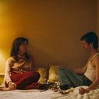 Ein Mann und eine Frau sitzen sich auf einem Bett gegenüber, in einem warm beleuchteten Raum mit einer Lampe links.