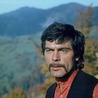 Ein Mann mit schwarzem Haar und Schnurrbart steht vor einer Berglandschaft. Er trägt ein rotes Hemd und eine schwarze Weste.