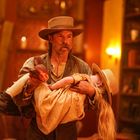 Ein Mann mit Hut hält ein bewusstloses Mädchen in einem hellen Raum. Bücherregale im Hintergrund.