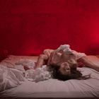 Rot beleuchtetes Schlafzimmer, Frau auf Bett, weiße Bettwäsche, rote Wand, Lampe rechts, Bücherstapel im Hintergrund links.