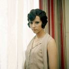 Eine Frau mit welligem, dunklem Haar trägt ein ärmelloses, beiges Kleid vor einem gestreiften Hintergrund.