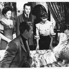 Fünf Menschen in einem Raum, einer liegt im Bett. Vier stehen, elegant gekleidet. Atmosphäre wirkt ernst und konzentriert.