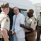 Ein Mann in weißem Anzug, umgeben von drei uniformierten Polizisten vor einem Fahrzeug mit Gitter.