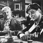 Zwei Männer sitzen an einem Tisch in einer Bar, die Wände sind mit vielen Bildern dekoriert. Links ein älterer, rechts junger Mann.