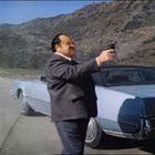 Ein Mann in einem Anzug steht auf einer Straße neben einem blauen Auto und zielt mit einer Pistole. Hintergrund: Hügel.
