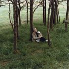 Keine Texte. Ein älterer Mann sitzt mit kleinem Kind auf einer Wiese im Wald, beide schauen nachdenklich.