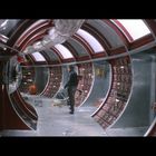 Keine sichtbaren Texte. Zylindrischer Raum mit roten Konsolen, Metallakzenten, Person in einem Raumanzug in futuristischer Umgebung.
