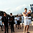 Person in weißer Sportkleidung hält eine Discus. Männer in Uniform und Anzügen im Hintergrund, Fahnen wehen.
