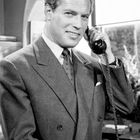 Ein Mann in einem klassischen Anzug telefoniert mit einem alten Telefon und lächelt leicht.