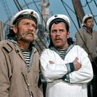 Zwei Männer in Matrosenkleidung und weißen Mützen auf einem Schiff. Sie schauen ernst in die Ferne.