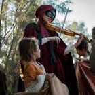 Eine Person mit rotem Umhang und Maske spielt Geige, umgeben von Kindern in historischen Kostümen unter Bäumen.