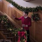Ein Mann in rotem Pullover steht vor einer weihnachtlich geschmückten Holztreppe, mit Lichterketten und Kränzen.