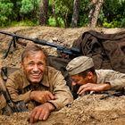 Ein lachender Mann und ein Soldat in Uniform liegend in einem Erdloch, umgeben von Bäumen und Ausrüstung.