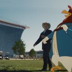 Ein großer cartoonhafter Hahn in blauem Anzug hält einen Golfschläger, während ein Mann im Anzug und Cowboyhut lacht.