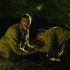 Zwei Personen mit blutverschmierten Gesichtern kauern in einem Wald, bei Nacht, und scheinen emotional aufgeladen zu sein.