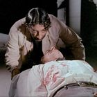 Zwei Männer in einem düsteren Raum. Einer liegt blutverschmiert am Boden, der andere kniet besorgt über ihm.