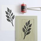 Eine schwarze Pflanzenillustration, links gedruckt auf weißem Papier, rechts der geschnitzte Stempel und eine Farbrolle.