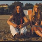 Text "Bagni Mosti" auf einem Gebäude. Zwei Personen sitzen am Strand, eine mit langen Haaren, die andere blond.