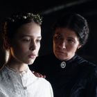 Eine junge Frau in Weiß mit Blumenkranz, daneben eine ältere Frau in dunkler Kleidung, die ihr die Hand auf die Schulter legt.