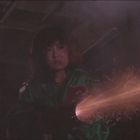 Unscharfe, rote Texttafeln im Hintergrund. Eine Person in grüner Jacke, hält eine Waffe abfeuernd mit Funken.