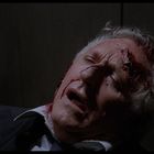 Kein Text erkennbar. Ein älterer Mann mit blutiger Stirn lehnt bewusstlos, Augen geschlossen, Mund leicht geöffnet an einer Wand.