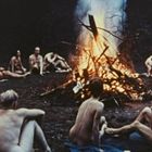 Kein sichtbarer Text. Menschen sitzen nackt im Kreis um ein großes Lagerfeuer im Freien.