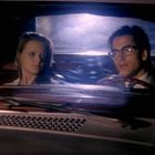 Ein Mann mit Brille und eine Frau sitzen im Auto. Es ist dunkel; Licht fällt durch die Windschutzscheibe ins Auto.