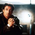 Ein Mann in dunkler Jacke sitzt nachdenklich, Hände gefaltet. Dahinter verschwommene Person auf Stuhl, dunkles Zimmer.