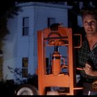 Eine Person in kariertem Hemd benutzt einen orangen Wagenheber. Hintergrund mit Haus bei Nacht.